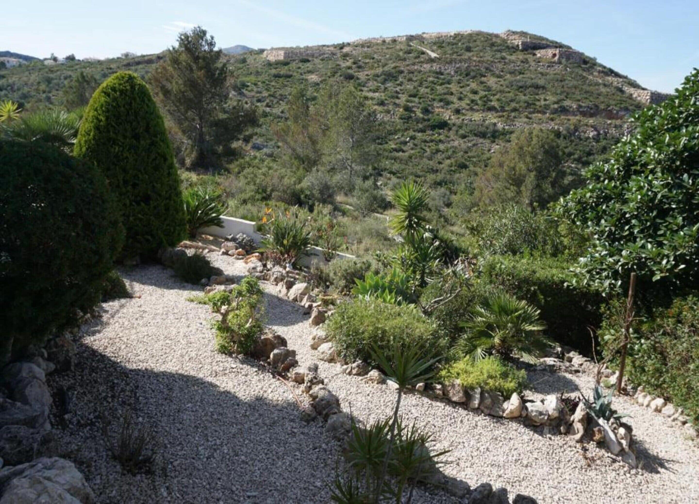 Un sendero de grava serpentea por un jardín ajardinado, con diversas plantas y una colina al fondo.