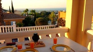 Mesa de comedor dispuesta con platos, cubiertos, una jarrón y vasos, con vistas a un paisaje que incluye edificios y árboles.