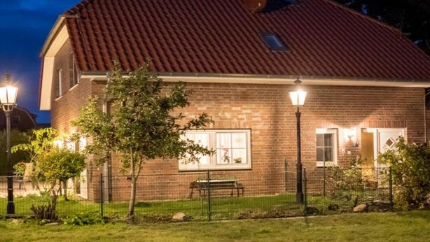 Una casa de ladrillo con techo de tejas rojas, un banco y un farol encendido en primer plano.