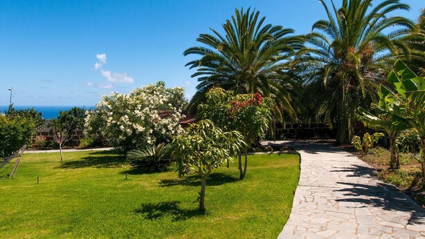A paved pathway curving through a well-maintained garden with palm trees and flowering shrubs, leading to a building with a red roof. - Villa in Gran Canaria With Private Pool & Tennis (Arucas)