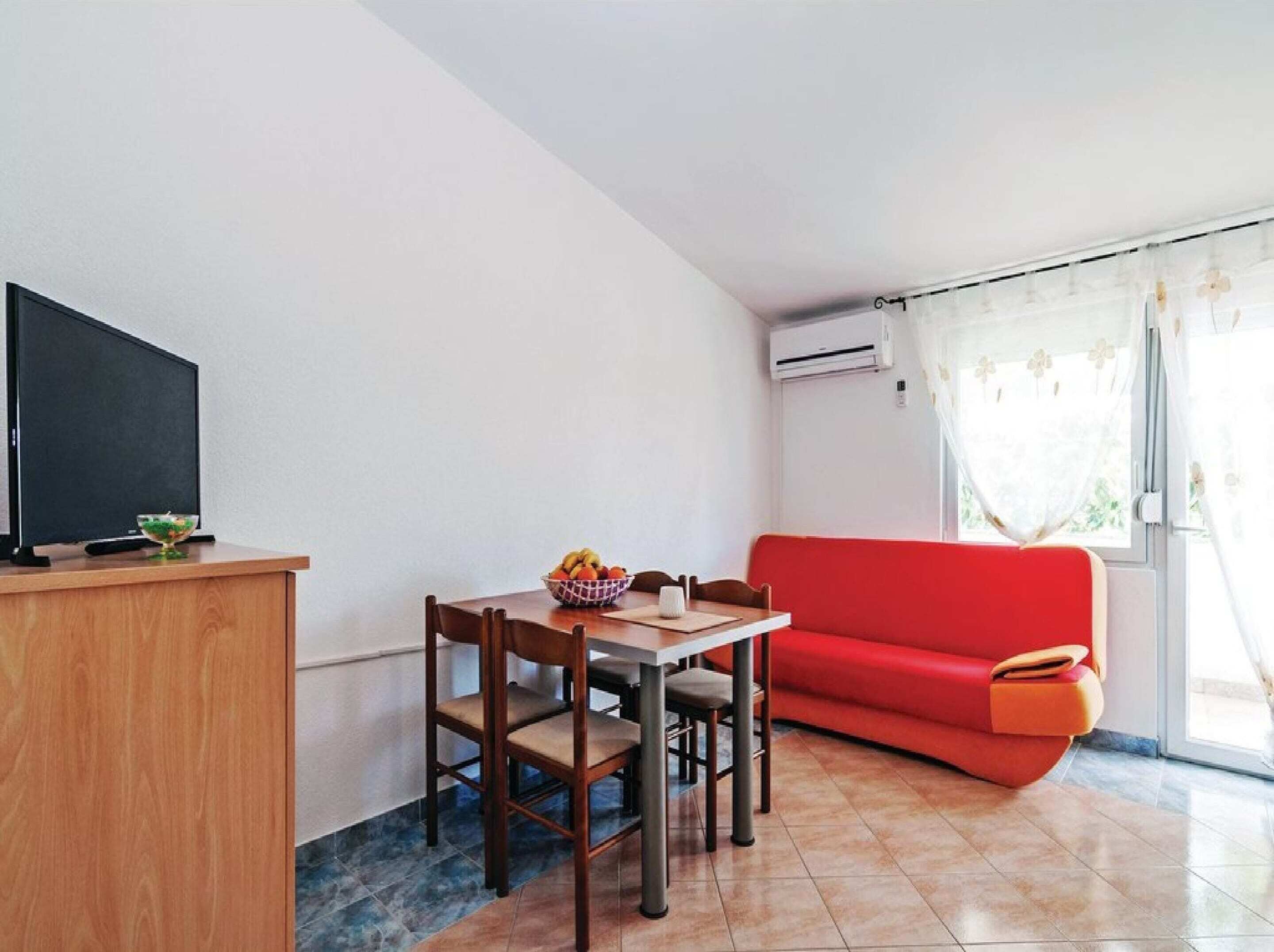 A compact living space with a dining table, two chairs, a red sofa, a TV on a wooden stand, and a window with curtains.