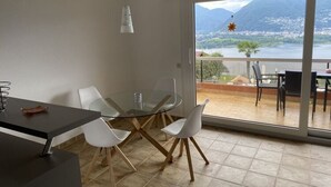 A modern dining area with a glass table and chairs, a black counter, and a view of a mountainous landscape through large windows. - Holiday Apartment Casa Chimp 5 (Gambarogno)