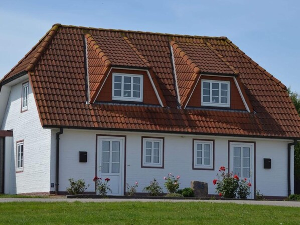 A two-story house with a brown tiled roof, white exterior walls, and a front garden with flowers. - South in Haus Luttmoor (Nordstrand)