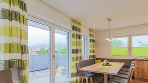 A modern dining area with a wooden table, chairs, and a bench. There is a sliding glass door leading to a balcony with a view of a green field and buildings. - Apartment in Brixen Near Ski Welt (Brixen im Thale)