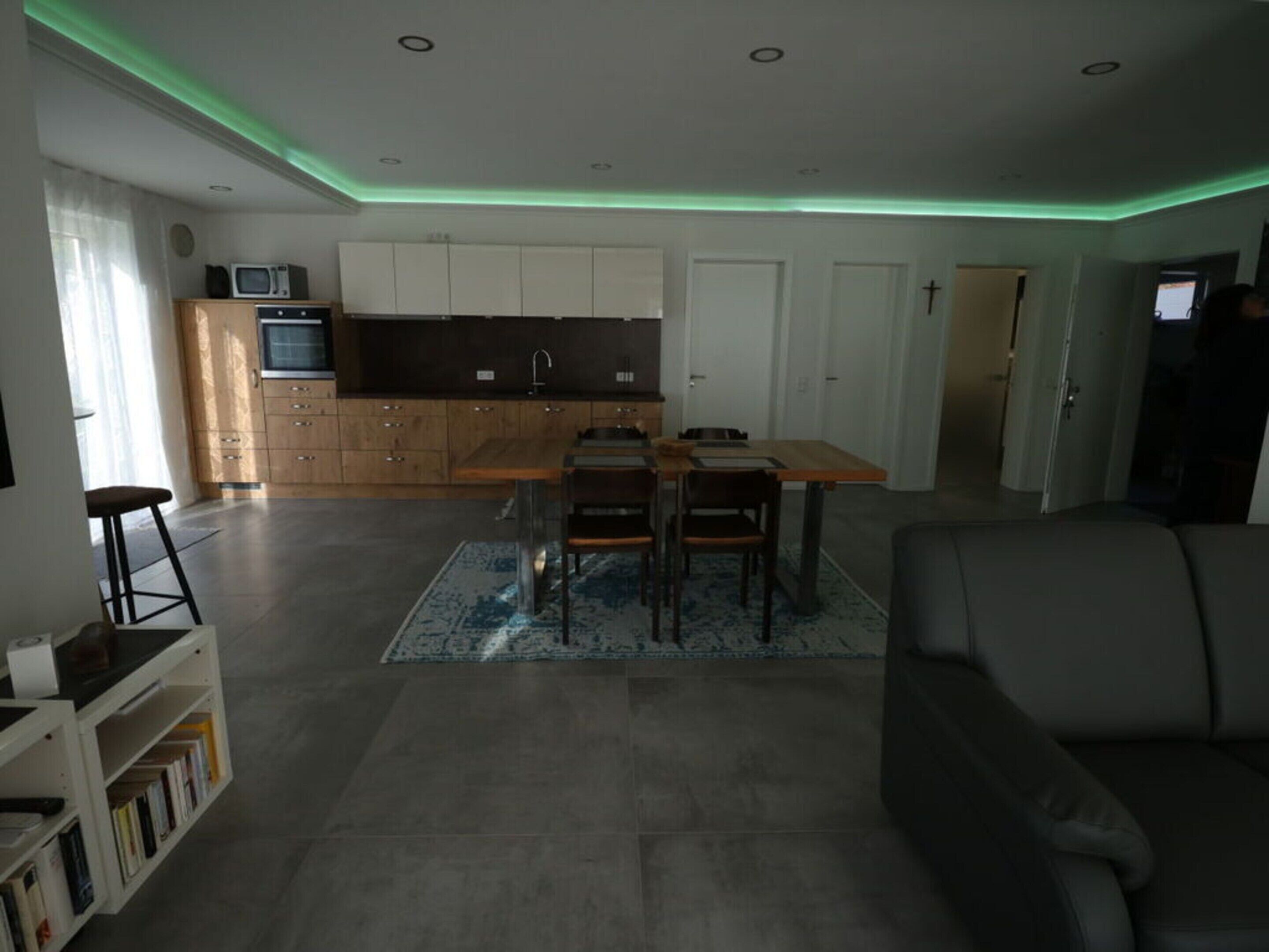 Una sala de estar moderna con un sofá gris, un comedor con mesa y sillas de madera, una cocina con armarios blancos y un microondas, y una pequeña estantería.