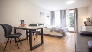 Una camera d'albergo moderna con pavimento in legno, un letto con coperta grigia, un tavolino con sedie, una scrivania con sedia e una finestra con tende.