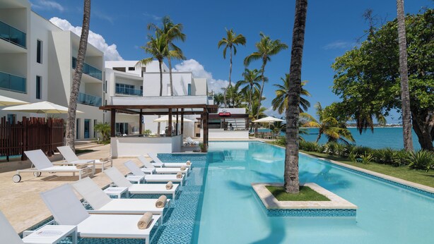 A pool area with lounge chairs and a covered seating area, surrounded by palm trees and a view of the ocean.