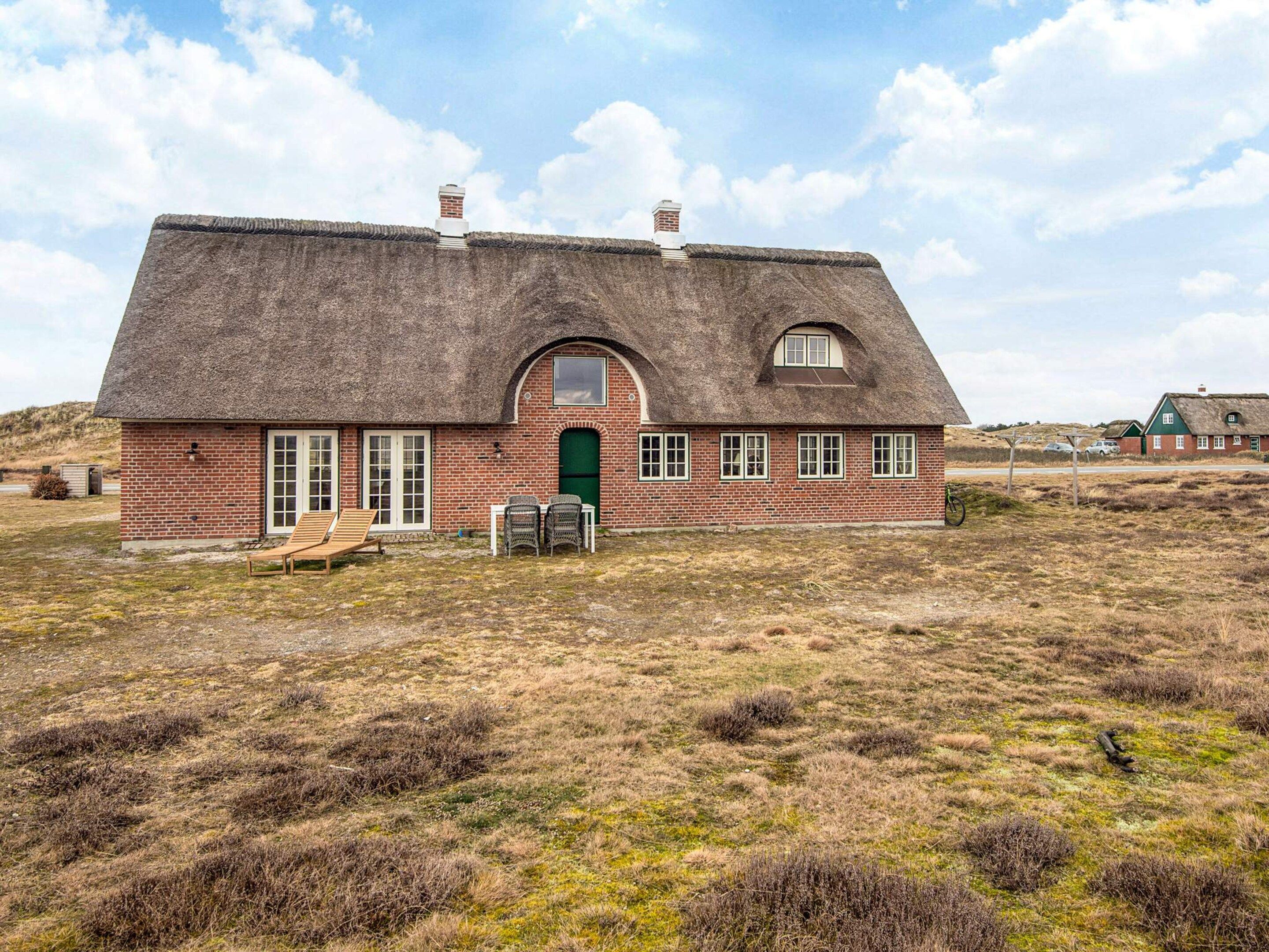 A thatched-roof house with a brick exterior, a green door, and a small porch with chairs.