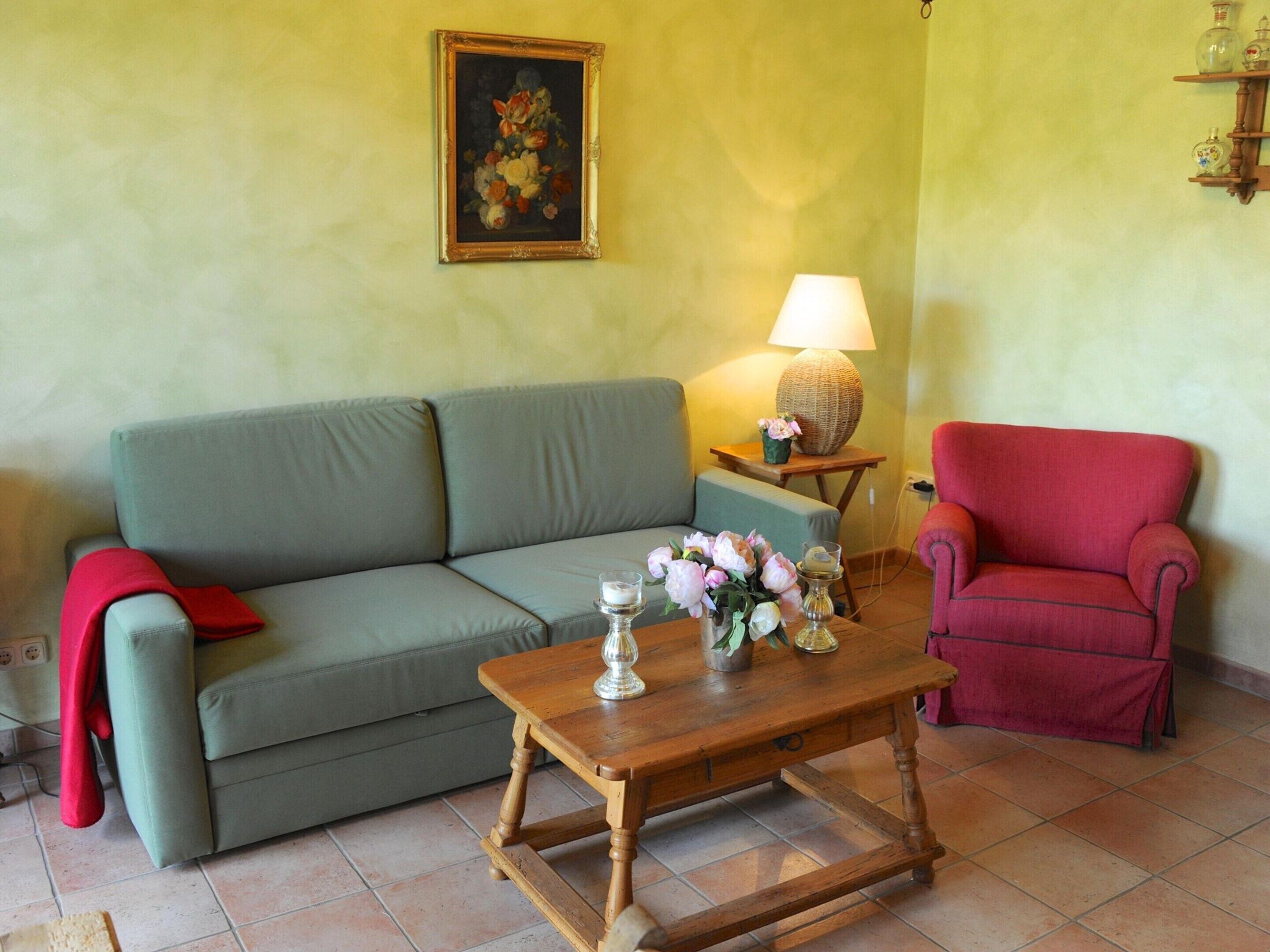 Una sala de estar con un sofá verde, una mesa de centro de madera con un jarrón de flores, un sillón rojo y una lámpara sobre una mesita auxiliar.