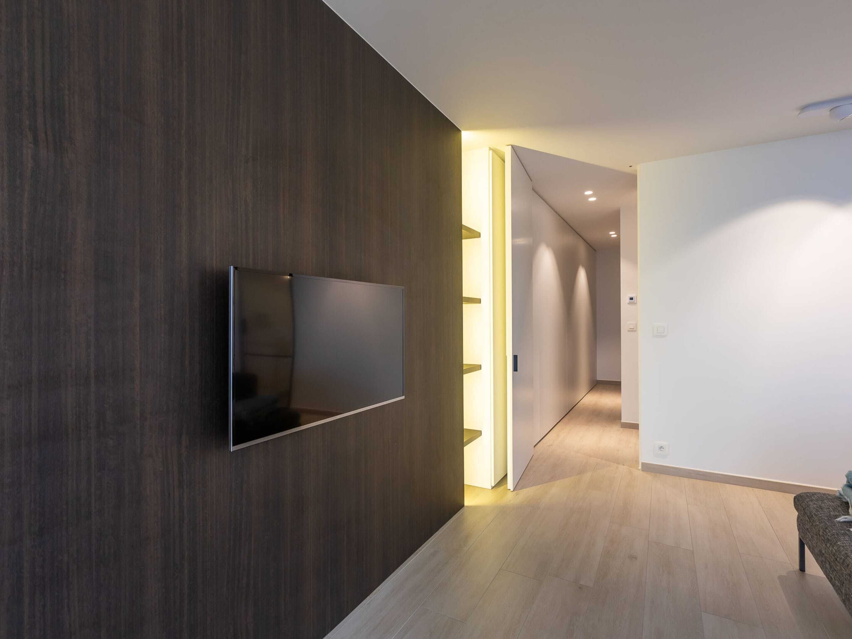 Ein moderner Flur mit einem an einer dunklen Holzwand befestigten Flachbildfernseher, der zu einem hellen Raum mit hellem Boden führt.