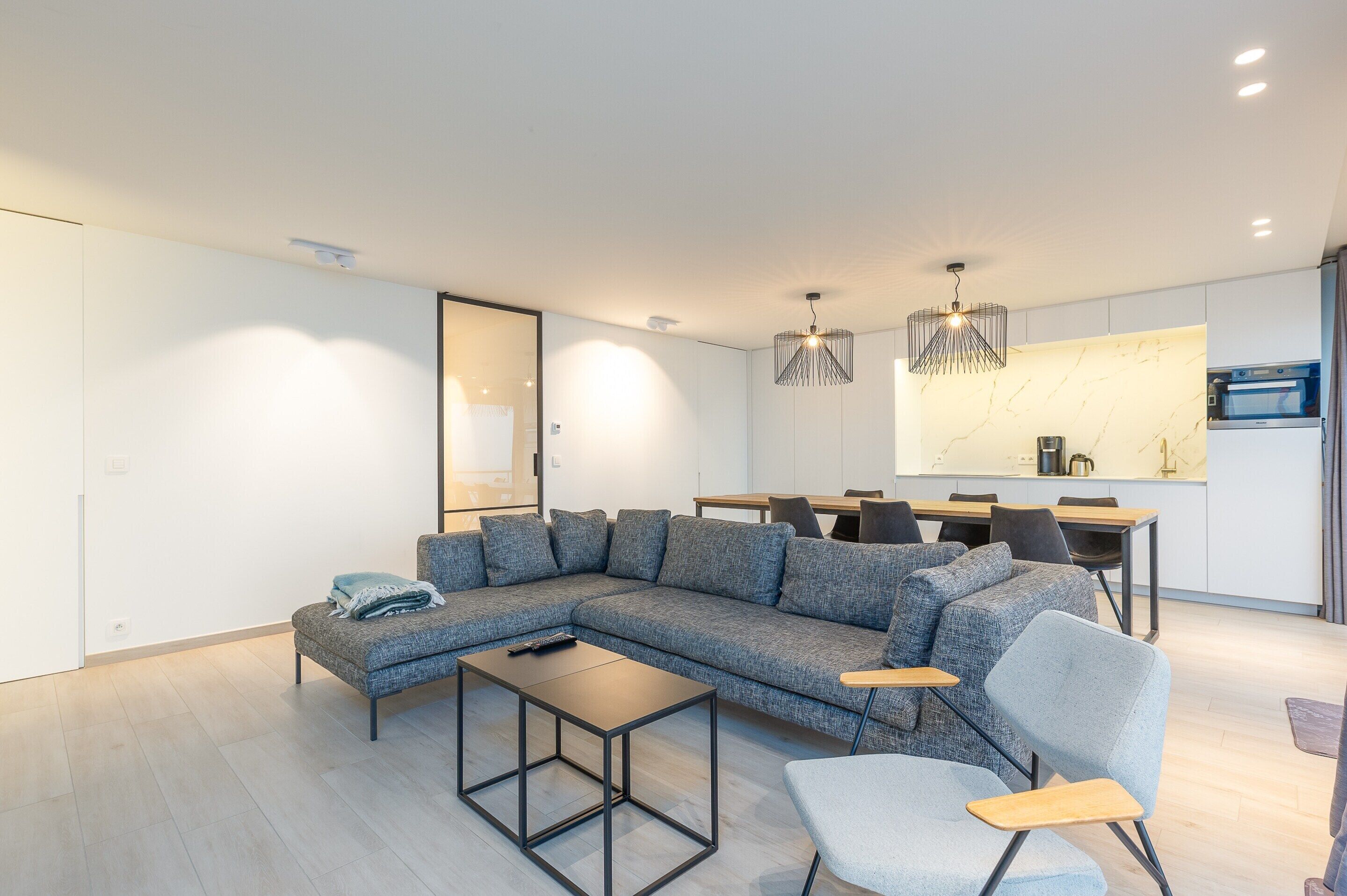 A modern living room with a sectional sofa, a coffee table, and a dining area with a kitchen in the background.