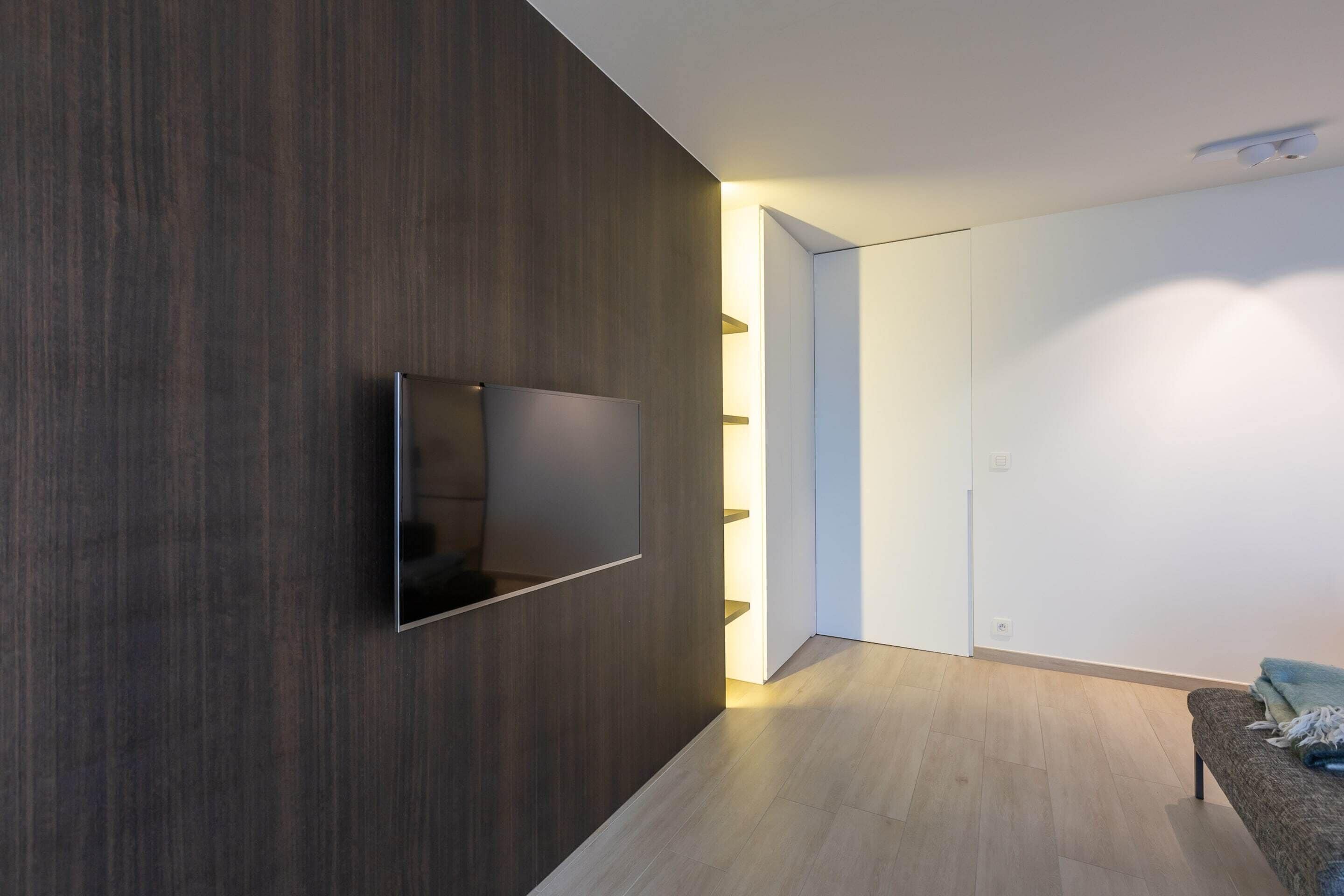 A modern room with a dark wooden accent wall, a flat-screen TV mounted on it, a light-colored wooden floor, and a sofa with a blanket.