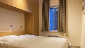 Un lit bien fait dans une chambre dotée d’une tête de lit en bois, d’une fenêtre avec des rideaux et d’une bouche d’aération fixée au mur.