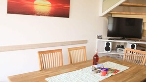 A dining area with a wooden table, chairs, a television, and a painting of a sunset. - Ostseebrandung - Haus Seestern (Dranske)