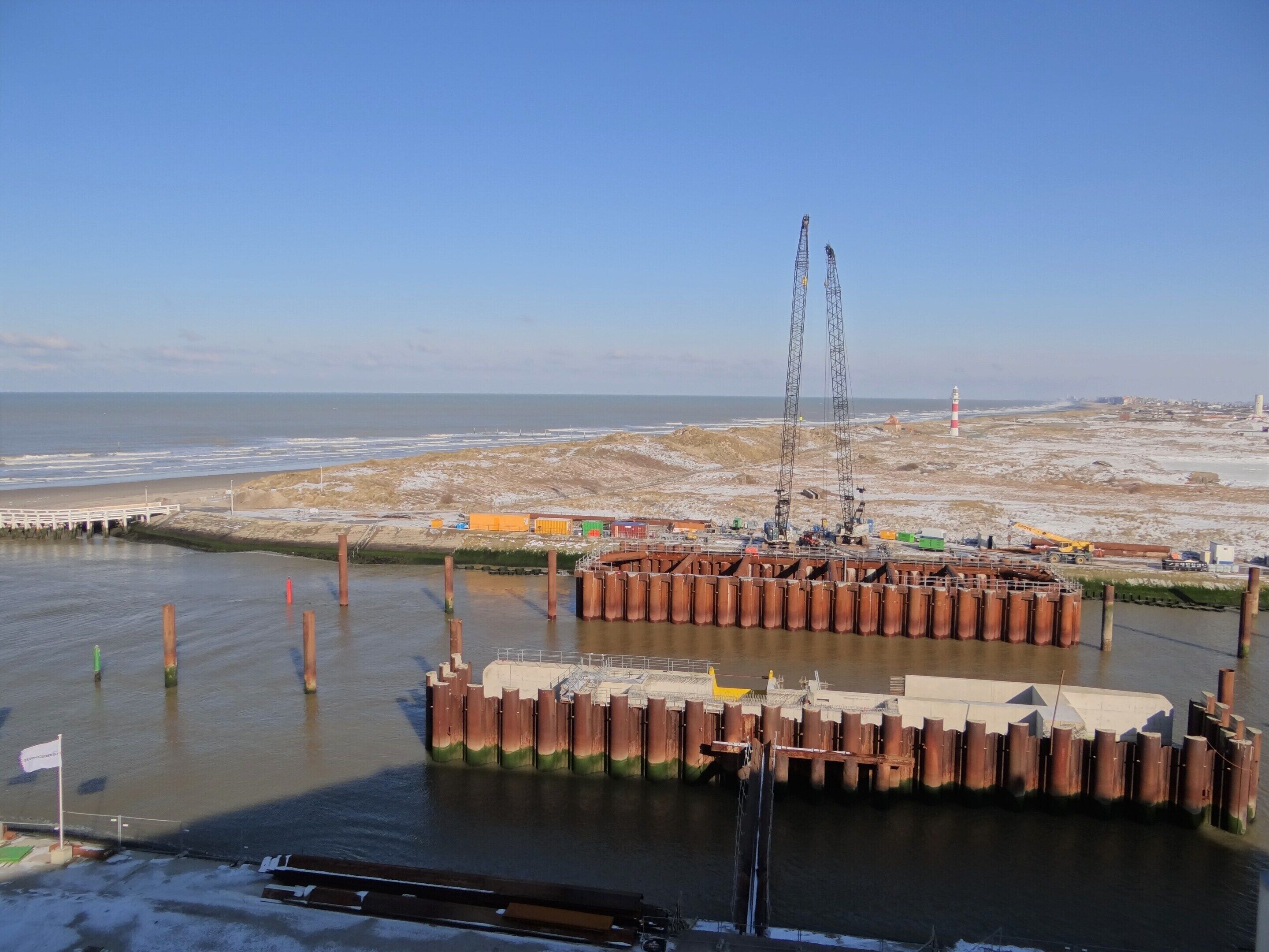 Un chantier de construction sur la côte, avec une grande grue, une digue et une plage de sable en arrière-plan.