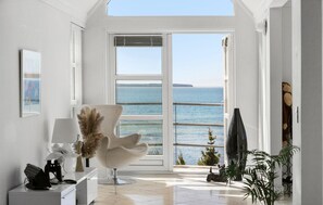 Una habitación moderna con un amplio ventanal con vista al mar, una silla cómoda, un escritorio con lámpara y una pequeña estatua de perro.