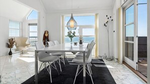 Een moderne eetruimte met een grote tafel en stoelen, een decoratieve hanglamp en uitzicht op zee door grote ramen.