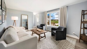 Ein Wohnzimmer mit einer Couch, zwei Sesseln, einem Couchtisch, einer Lampe, einem Spiegel und Blick auf eine grüne Landschaft durch ein Fenster.