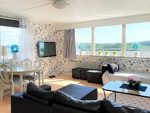 Un salon comprenant un coin repas, un téléviseur à écran plat fixé au mur et une grande fenêtre donnant sur un paysage verdoyant.