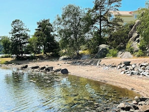 A clear view of a beach with rocks, trees, and a building in the background. - 4 Star Holiday Home in Uddevalla (Uddevalla)