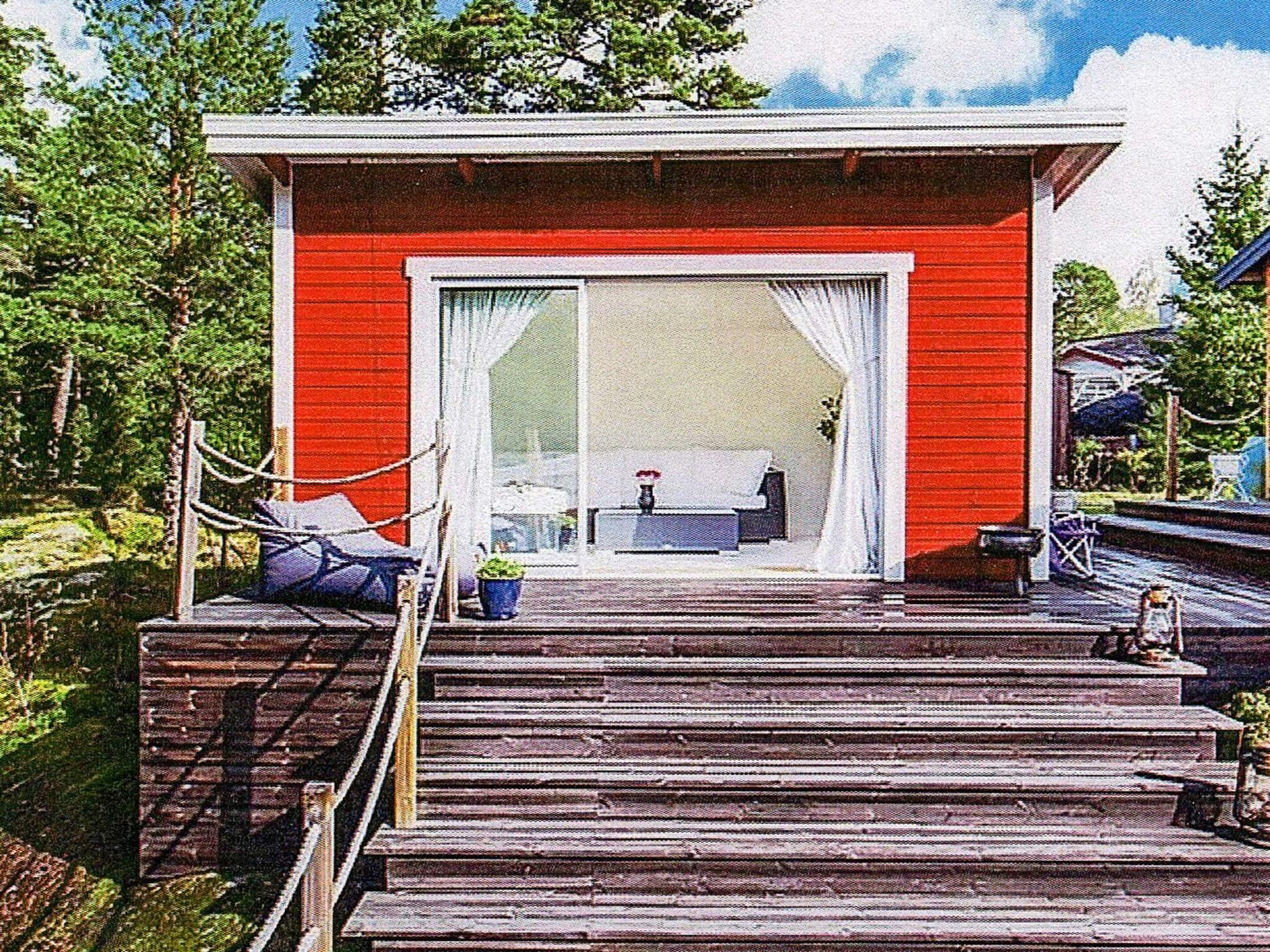 A red building with a large sliding glass door, a wooden deck leading to a seating area with a table and chairs, and a potted plant.