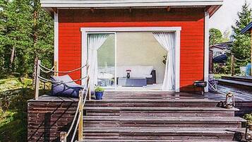 A red building with a large sliding glass door, a wooden deck leading to a seating area with a table and chairs, and a potted plant.