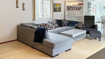 Ein modernes Wohnzimmer mit einem grauen Ecksofa, einem Couchtisch, einem Bücherregal und einem Essbereich mit Tisch und Stühlen.