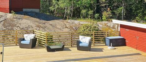 Un espace aménagé au bord de la piscine, avec une terrasse en bois, deux fauteuils en osier dotés de coussins, une petite table et, au fond, un bâtiment rouge.