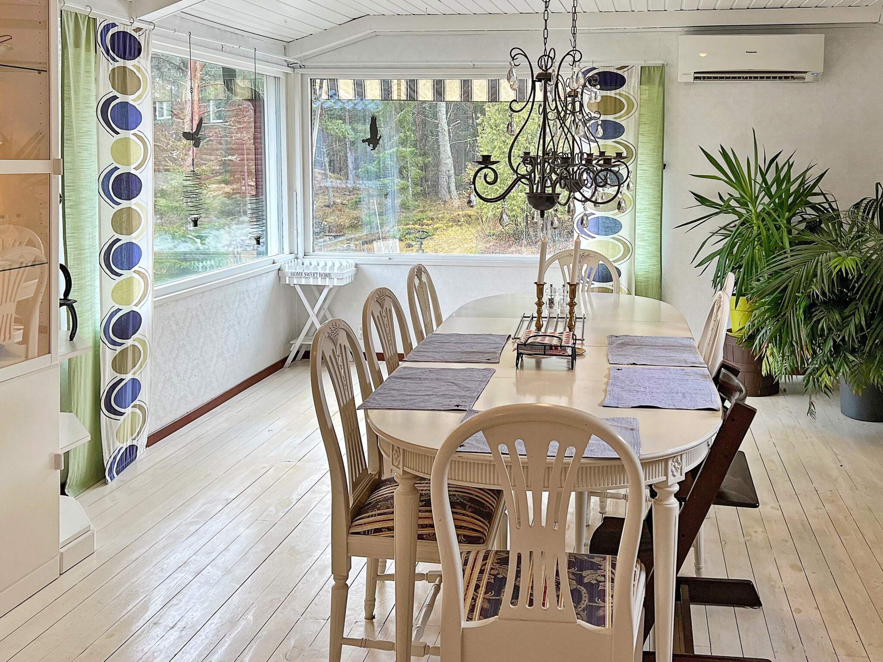 A dining area with a long table, chairs, a chandelier, and a view of trees through large windows.