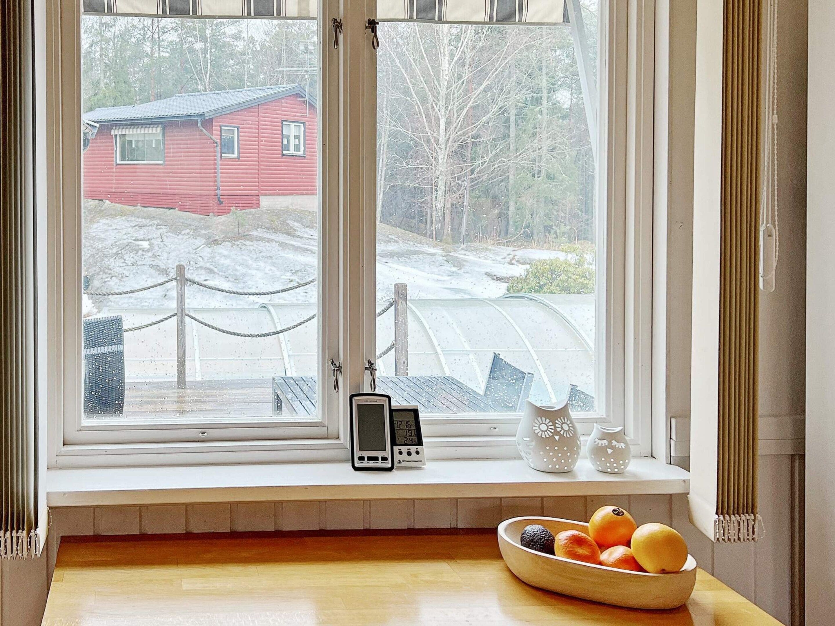 Une pièce avec une fenêtre donnant sur une maison rouge et un paysage enneigé, une table en bois sur laquelle se trouve un saladier rempli de fruits, et un thermostat fixé au mur.