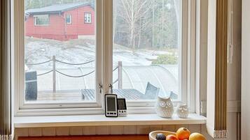Une pièce avec une fenêtre donnant sur une maison rouge et un paysage enneigé, une table en bois sur laquelle se trouve un saladier rempli de fruits, et un thermostat fixé au mur.
