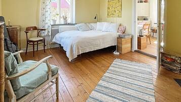 Un dormitorio con cama, una silla, una mesita de noche, lámpara, ventana con cortinas y puerta que da a otra habitación.