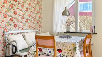 Una habitación acogedora con papel tapiz floral, una cama con cabecera blanca, una silla de madera, una mesa con mantel floreado, una lámpara colgante y una ventana con vistas a árboles.