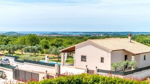 Una casa moderna con techo de tejas, un porche cubierto y vistas a un paisaje verde y agua al fondo.