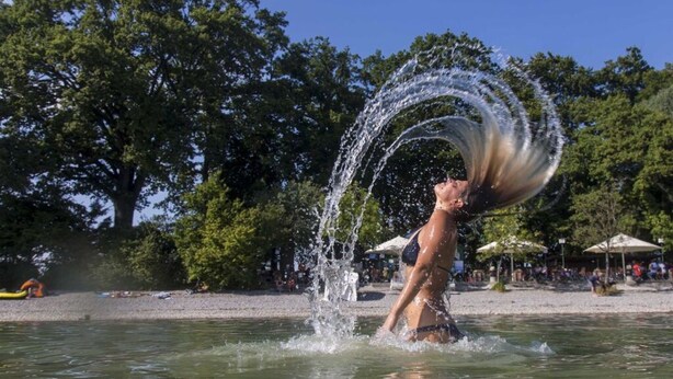 Una persona en traje de baño jugando con agua en una playa con árboles y sombrillas al fondo.