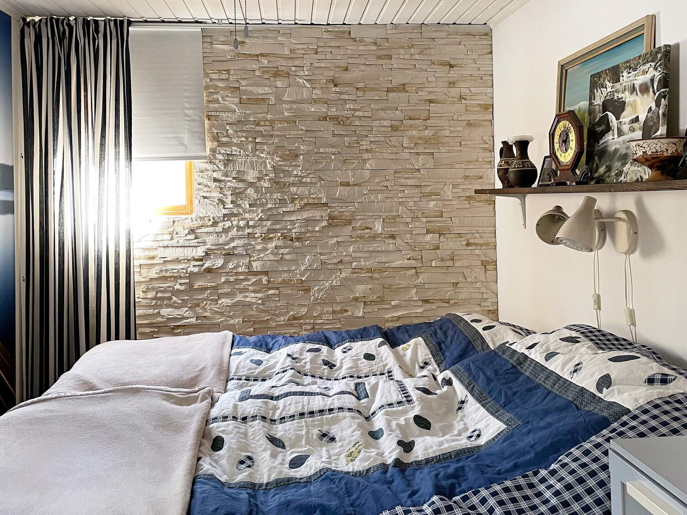Ein Schlafzimmer mit Steinwand, einem Bett mit blau-weißer Steudecke, einer Nachttischlampe und einer Uhr an der Wand.