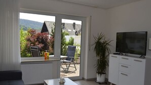 A living room with a sofa, a coffee table, a TV, and a sliding glass door leading to a patio with a chair and plants. - Palatinate Forest Comfortable Holiday Residence (Silz)