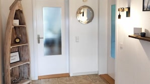 Un pasillo con una puerta blanca, un estante de madera con un reloj, un espejo y un pequeño estante con una taza y un letrero.