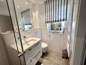 A modern bathroom with a glass shower enclosure, a white sink with a mirror, a toilet, and a window with striped curtains. - Holiday Home Sonnenhus (St. Peter-Ording)