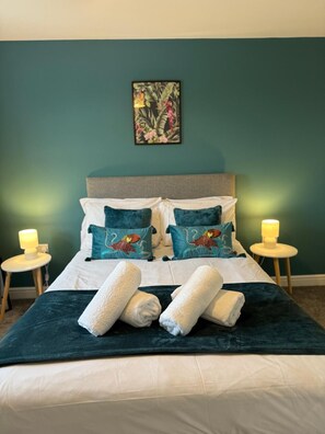 A neatly made bed with two pillows, a decorative pillow with a bird design, and two rolled towels. There are two bedside lamps on wooden nightstands. - Charming 5BR Hawarden Sleeps 10 | Contractor Stays (Hawarden)