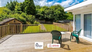 A wooden deck with a view of a garden and a house, a table with chairs, and logos for Harper and Airbnb. - 4BR Home | Sleeps 10 | 7 Beds for Family & Crew (Flintshire)