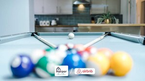 A pool table with a white cue ball and colorful balls, a kitchen area with cabinets and a plant, and logos for Harper and Airbnb. - 4BR Home | Sleeps 10 | 7 Beds for Family & Crew (Flintshire)