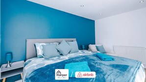 A modern bedroom with a blue wall, a bed with a patterned bedspread, and a nightstand with a lamp. - 4BR Home | Sleeps 10 | 7 Beds for Family & Crew (Flintshire)