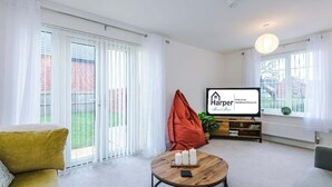 A modern living room with a sofa, a coffee table with candles, a television, and a view of a garden through large windows. - 4BR Home | Sleeps 10 | 7 Beds for Family & Crew (Flintshire)