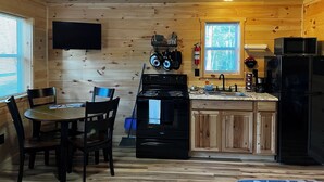 Een keuken met houten lambrisering, voorzien van een fornuis, magnetron en koelkast. Er is een eettafel met stoelen, een raam en een flatscreen televisie aan de wand bevestigd.