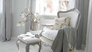 A room with a white sofa, a small table with a vase of flowers, and a window with curtains. - Liefde Voor de Schlei (Arnis)