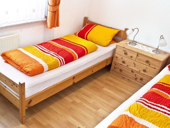 A small, single-bed room with a wooden frame, a white mattress, and a striped blanket. There is a wooden nightstand with two lamps and a drawer. - A in Haus Seewind Comfortable Vacation Home (Wittduen)