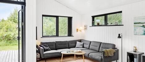 Un salon moderne avec un canapé d’angle gris, une petite table basse et une cheminée. De grandes fenêtres laissent entrer la lumière naturelle et offrent une vue sur l’extérieur.