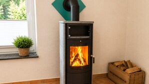 Un poêle à bois installé dans un coin, près d’une fenêtre, avec une petite plante et une boîte de bois de chauffage.