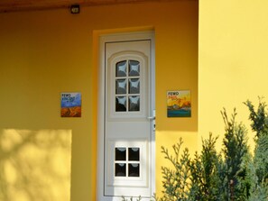 Ein gelbes Gebäude mit einer weißen Tür, zwei gerahmten Bildern an der Wand und einer Pflanze im Vordergrund.